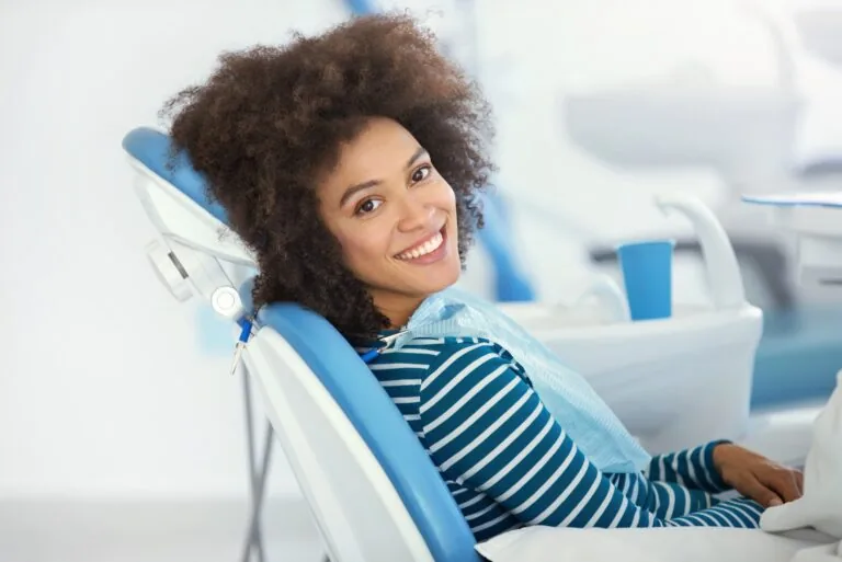 Patient sitting in a dental chair at a dentist's office, smiling, wearing a striped shirt and dental bib. Dental equipment and instruments are visible in the clinical setting.