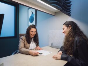 Two individuals sit across from each other at a desk in a modern office with a privacy partition and abstract blue artwork. One points to a document with a pen while the other listens attentively, suggesting a professional consultation or advisory session.