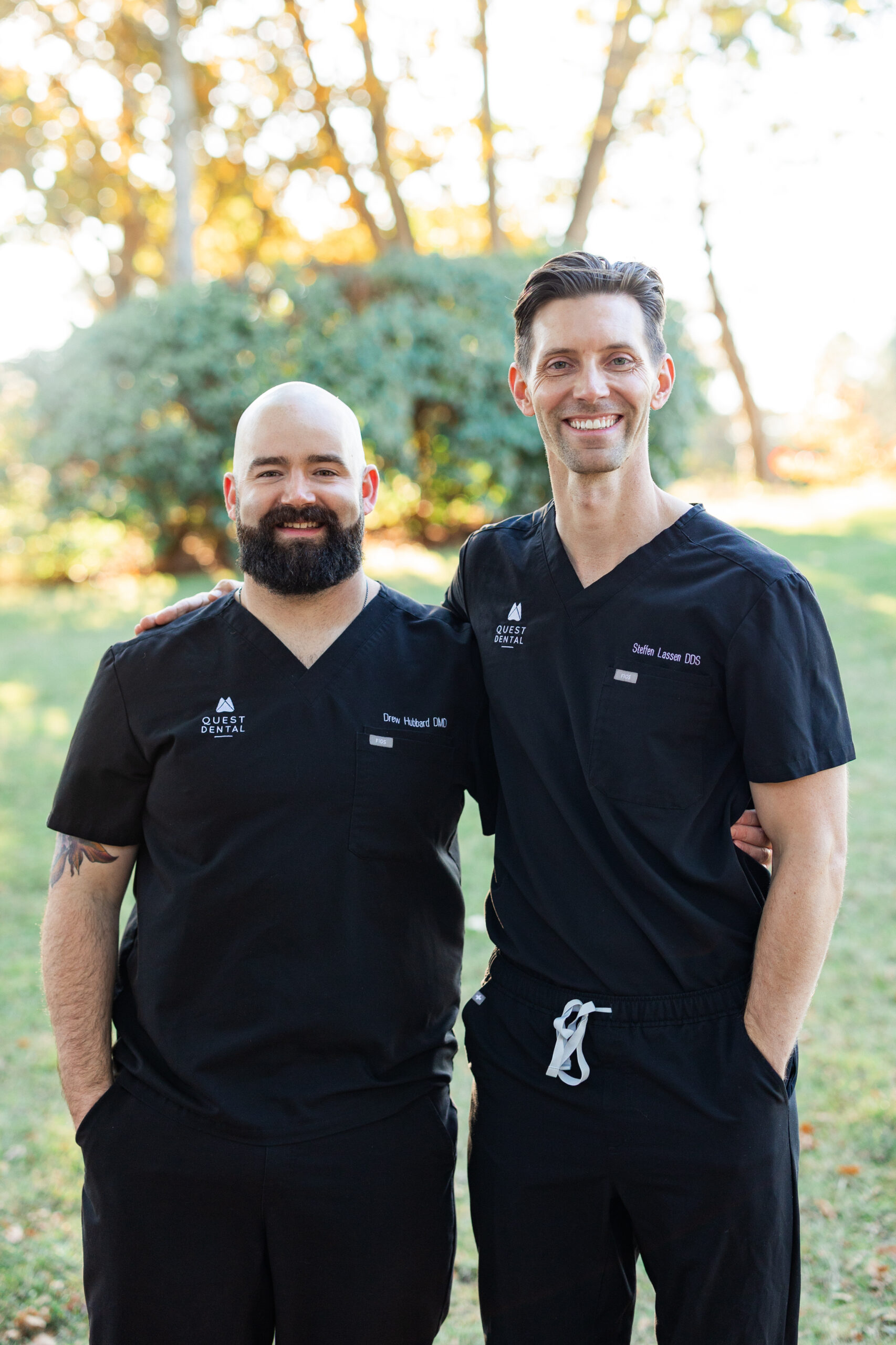 Two individuals in black scrubs stand outdoors on grass with trees behind them. They smile warmly, with one placing an arm around the other, suggesting camaraderie in a professional setting.