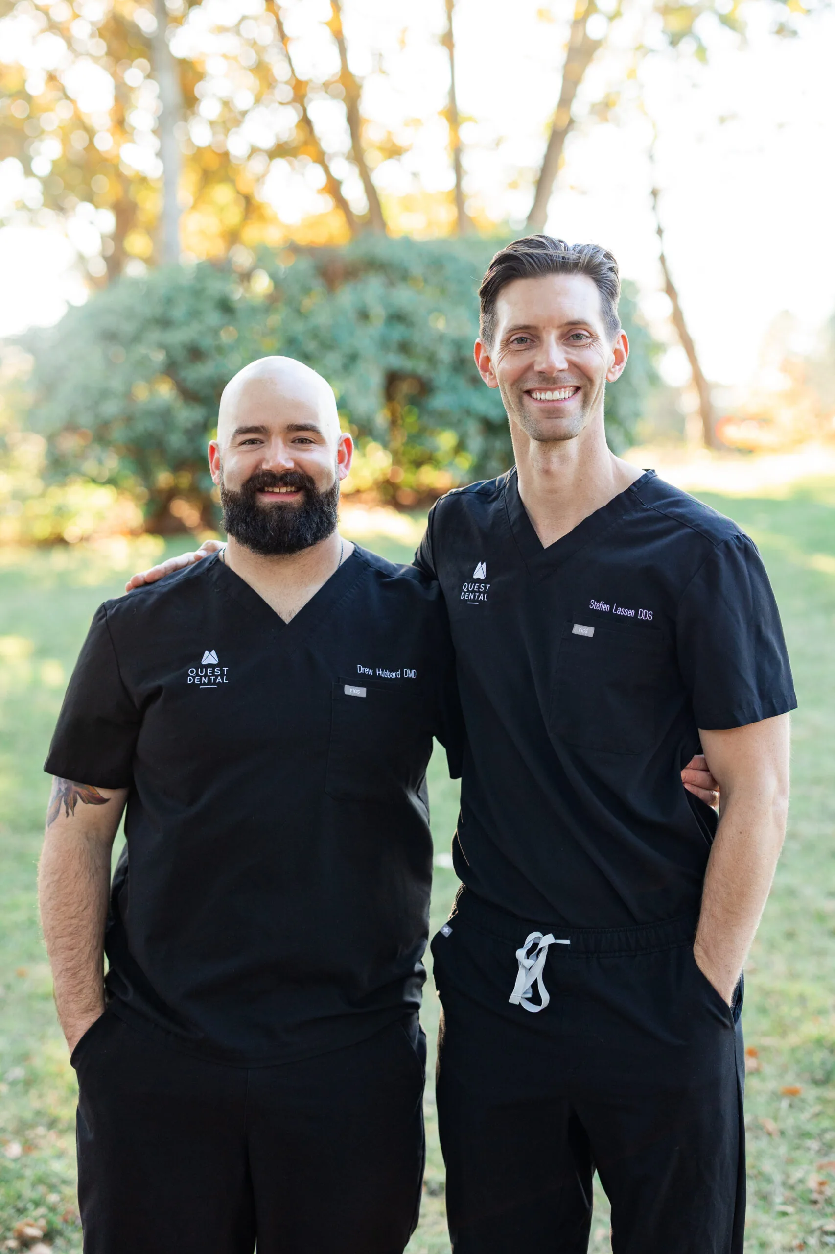 Two individuals in black scrubs stand outdoors on grass with trees behind them. They smile warmly, with one placing an arm around the other, suggesting camaraderie in a professional setting.