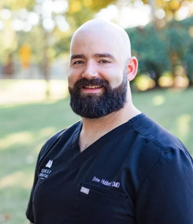 Dr. Hubbard in black scrubs embroidered with “QUEST DENTAL” and “Drew Hubbard DMD” stands outdoors, smiling warmly. The park-like setting with trees and grass creates a natural, approachable atmosphere for this professional portrait.