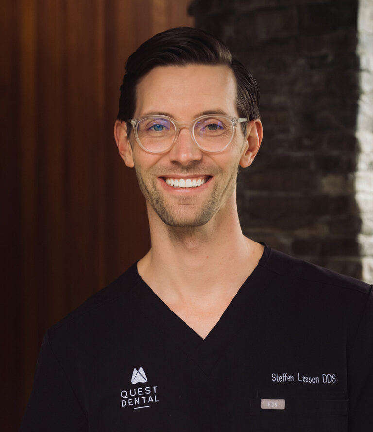 Person wearing black scrubs embroidered with “Steffen Lassen DDS” and “QUEST DENTAL,” smiling in front of a dark stone and wood-paneled wall. Clear-framed glasses and the indoor setting suggest a professional portrait in a dental office.