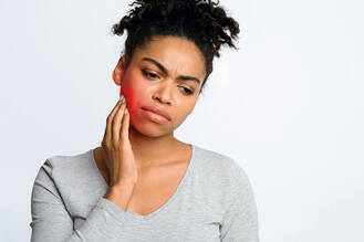 A woman with curly hair in a gray top is holding her cheek, highlighted red, showing tooth or jaw pain.